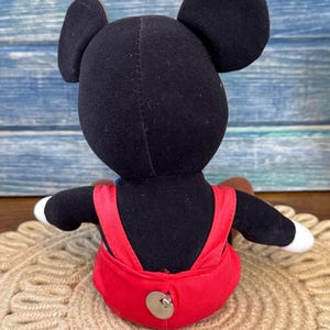 May include: A plush Mickey Mouse doll, seen from the rear, with black ears, a black body, and red dungarees. The doll has white gloves and a brown tail. It is sitting on a beige, circular woven mat.