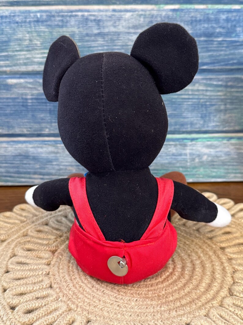 May include: A plush Mickey Mouse doll, seen from the rear, with black ears, a black body, and red dungarees. The doll has white gloves and a brown tail. It is sitting on a beige, circular woven mat.