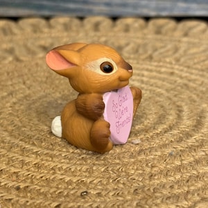 May include: A brown bunny figurine holding a pink heart-shaped sign. The sign reads "So Glad We're Friends!" The bunny has a white tail, pink ears, and black eyes. The figurine is sitting on a woven, tan surface.