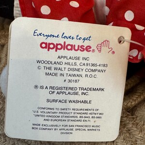 May include: A product tag from Applause Inc. is attached to a red and white polka dot fabric. The tag includes the text "Everyone loves to get applause." and "Surface Washable."