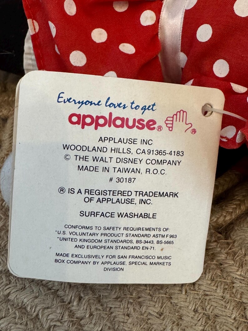 May include: A product tag from Applause Inc. is attached to a red and white polka dot fabric. The tag includes the text "Everyone loves to get applause." and "Surface Washable."