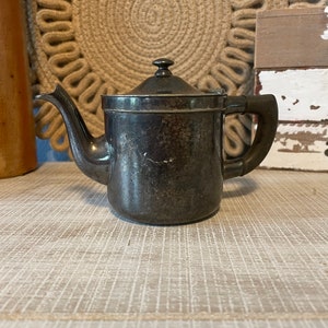 May include: A silver teapot with a tarnished finish and a black handle. The teapot has a rounded body and a spout on the side.