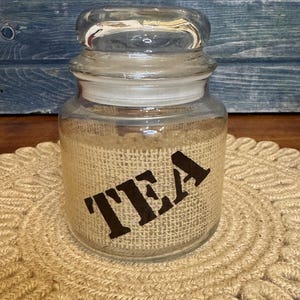 May include: A clear glass jar with a lid, featuring a burlap wrap with the word "TEA" in dark brown letters. The jar sits on a round, woven, beige placemat. The background is a blue wooden surface.