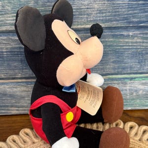 May include: A plush Mickey Mouse toy, with black ears, a black body, and a tan face. Mickey wears red overalls with a yellow button and a blue bow tie. The toy has brown feet and a small tag.