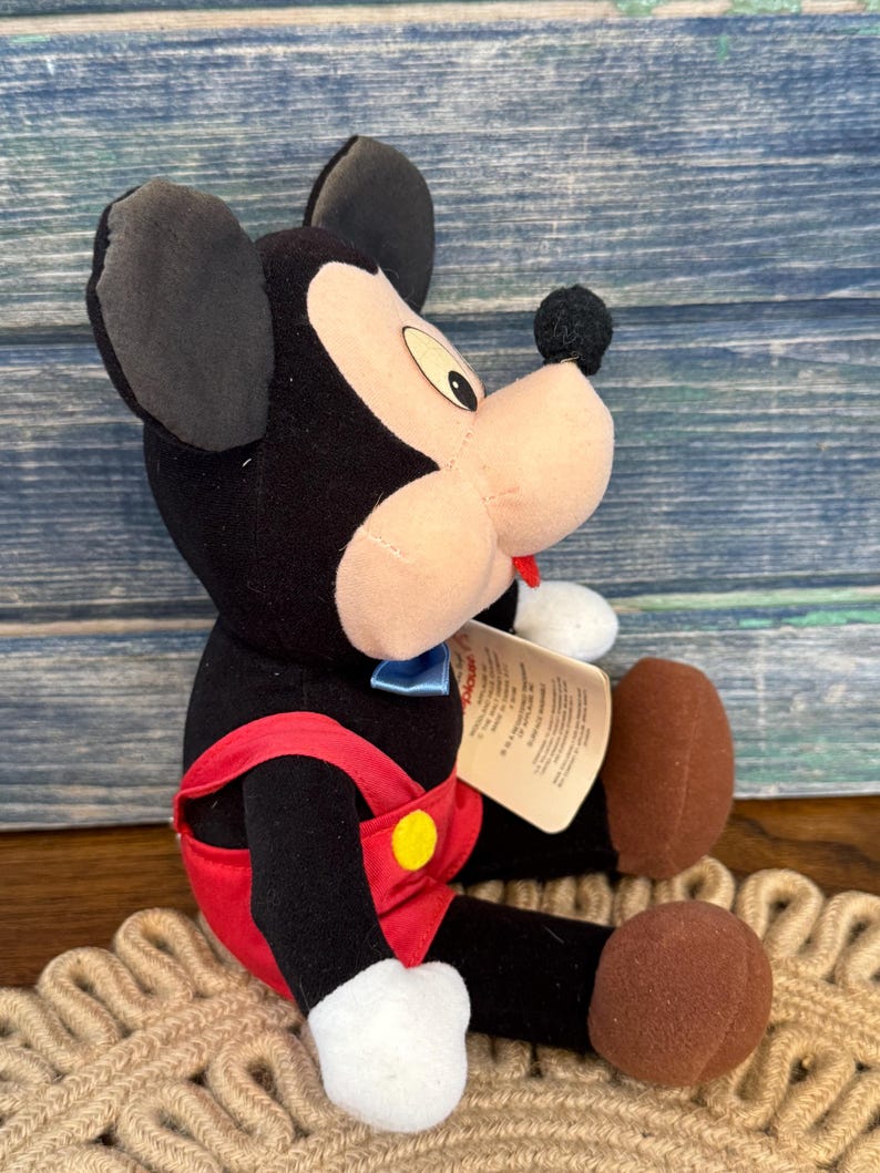 May include: A plush Mickey Mouse toy, with black ears, a black body, and a tan face. Mickey wears red overalls with a yellow button and a blue bow tie. The toy has brown feet and a small tag.