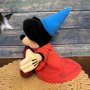 Puede incluir: Un peluche de Mickey Mouse vestido de hechicero, con un sombrero azul y una t&uacute;nica roja. El juguete tiene orejas negras, una cara negra y guantes blancos con detalles negros. Est&aacute; sentado sobre una alfombra redonda tejida.