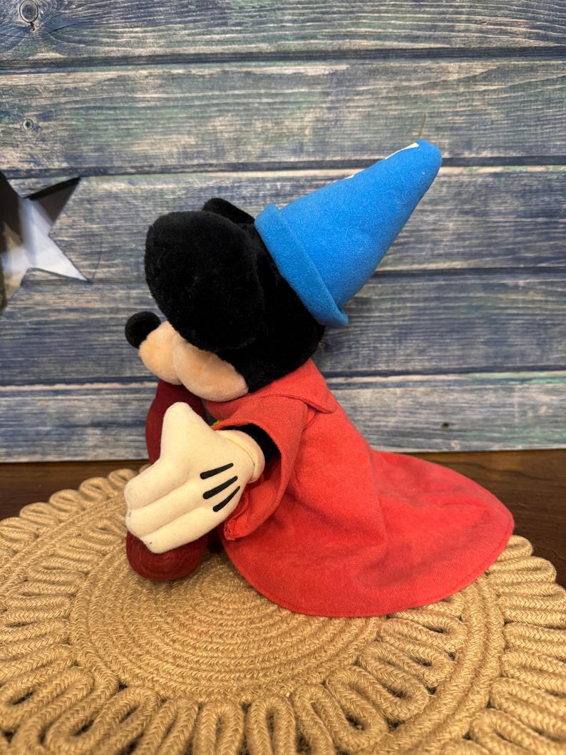 Puede incluir: Un peluche de Mickey Mouse vestido de hechicero, con un sombrero azul y una t&uacute;nica roja. El juguete tiene orejas negras, una cara negra y guantes blancos con detalles negros. Est&aacute; sentado sobre una alfombra redonda tejida.