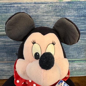 May include: A Minnie Mouse soft toy featuring black ears, a black nose, and a red and white polka dot bow. The doll has a tag attached. This plush toy is a classic Disney character.