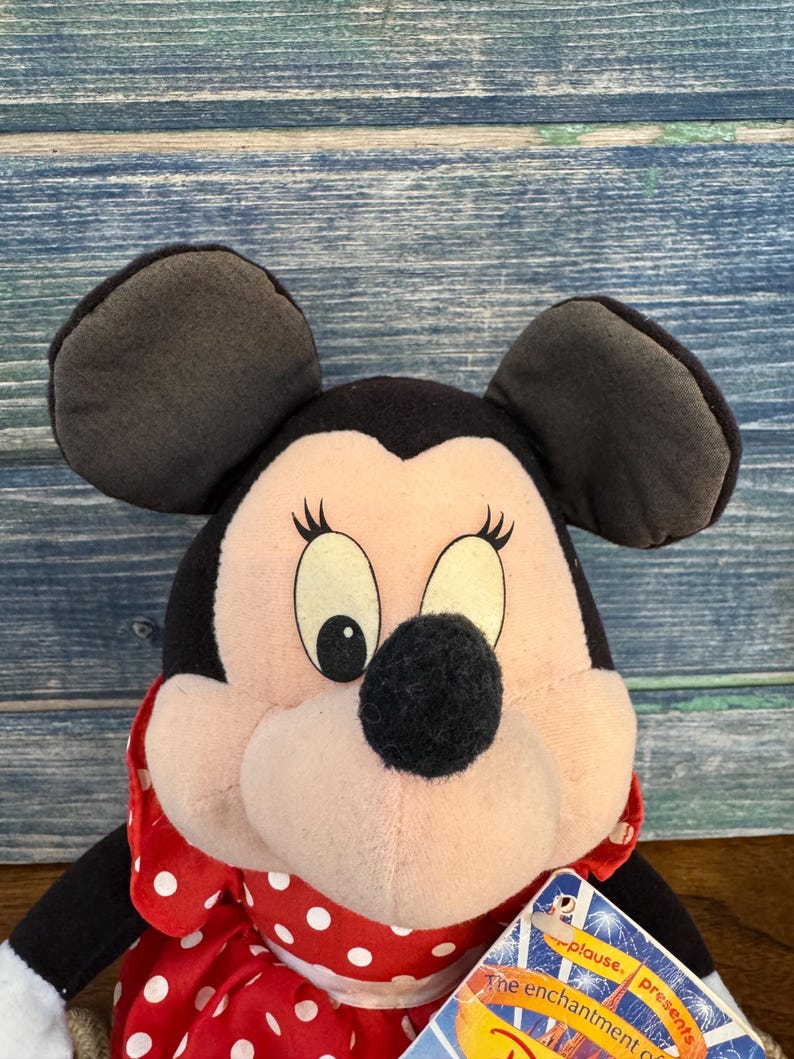 May include: A Minnie Mouse soft toy featuring black ears, a black nose, and a red and white polka dot bow. The doll has a tag attached. This plush toy is a classic Disney character.