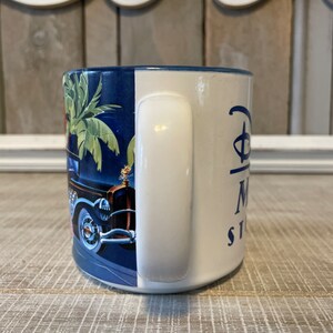 Puede incluir: Una taza de cer&aacute;mica azul y blanca con un dise&ntilde;o de coche antiguo y palmeras. La taza tiene un asa blanca y el texto "Disney's Magic Kingdom" en el lateral.