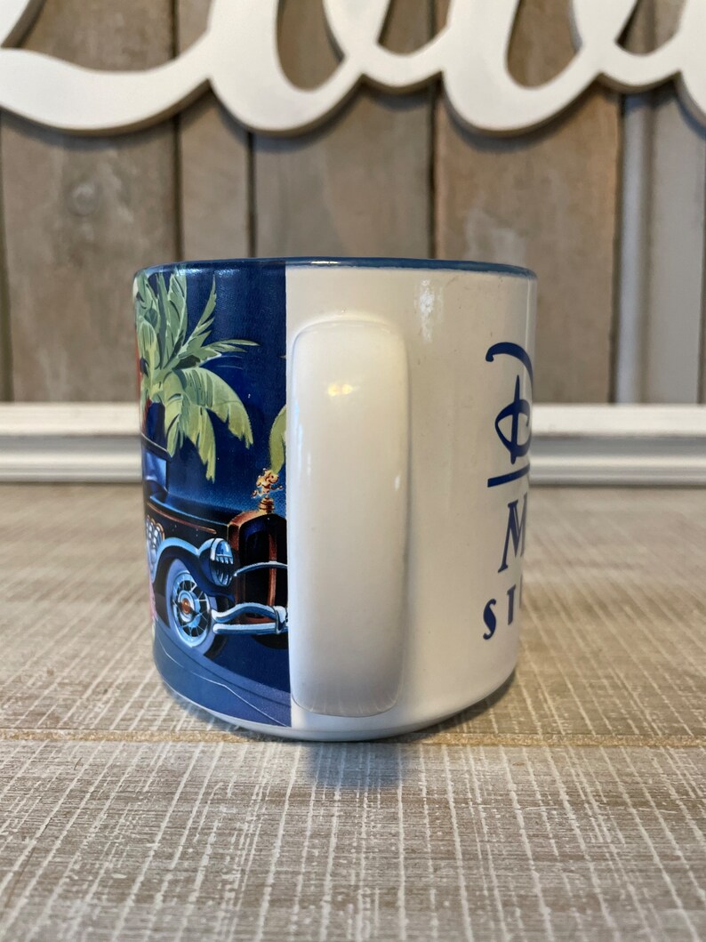 Puede incluir: Una taza de cer&aacute;mica azul y blanca con un dise&ntilde;o de coche antiguo y palmeras. La taza tiene un asa blanca y el texto "Disney's Magic Kingdom" en el lateral.
