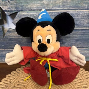 Puede incluir: Un peluche de Mickey Mouse vestido de hechicero, con una t&uacute;nica roja, zapatos rojos y un sombrero azul con una luna. El juguete tiene orejas negras, guantes blancos y una cuerda amarilla. Est&aacute; sobre una estera tejida.