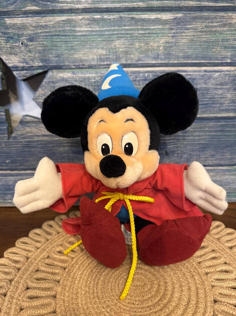 Puede incluir: Un peluche de Mickey Mouse vestido de hechicero, con una t&uacute;nica roja, zapatos rojos y un sombrero azul con una luna. El juguete tiene orejas negras, guantes blancos y una cuerda amarilla. Est&aacute; sobre una estera tejida.