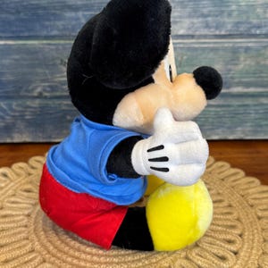 Peut inclure: Une peluche Mickey Mouse, avec une t&ecirc;te et des oreilles noires, un visage beige et une chemise bleue. Le jouet a un short rouge, des pieds jaunes et des gants blancs avec des lignes noires. Le jouet est assis sur un tapis tiss&eacute; marron.