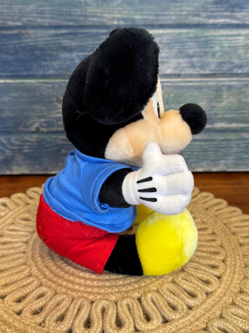 Peut inclure: Une peluche Mickey Mouse, avec une t&ecirc;te et des oreilles noires, un visage beige et une chemise bleue. Le jouet a un short rouge, des pieds jaunes et des gants blancs avec des lignes noires. Le jouet est assis sur un tapis tiss&eacute; marron.