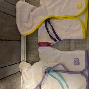 May include: Two white hooded towels with colorful trim. The top towel has yellow, pink, and purple trim. The bottom towel has purple, pink, and blue trim. Both towels have a small logo on the front.