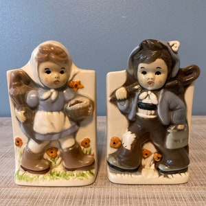 May include: A pair of vintage ceramic bookends featuring a girl and a boy. The girl is wearing a blue dress and a brown hat, and she is holding a basket. The boy is wearing a blue jacket and a brown hat, and he is holding a stick. Both bookends have a white background with orange flowers.