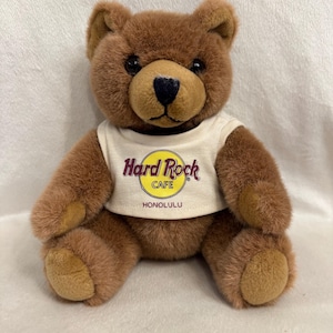 May include: A brown teddy bear wearing a white t-shirt with the Hard Rock Cafe logo and the word "HONOLULU" underneath. The bear has a black nose and dark eyes. The plush toy is sitting upright.