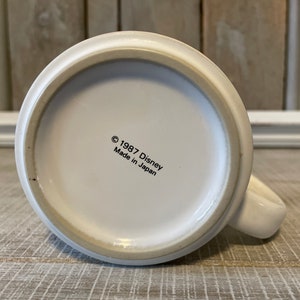 Puede incluir: Taza de cer&aacute;mica blanca con asa negra. En la parte inferior de la taza aparece el texto "&copy; 1987 Disney Hecho en Jap&oacute;n".