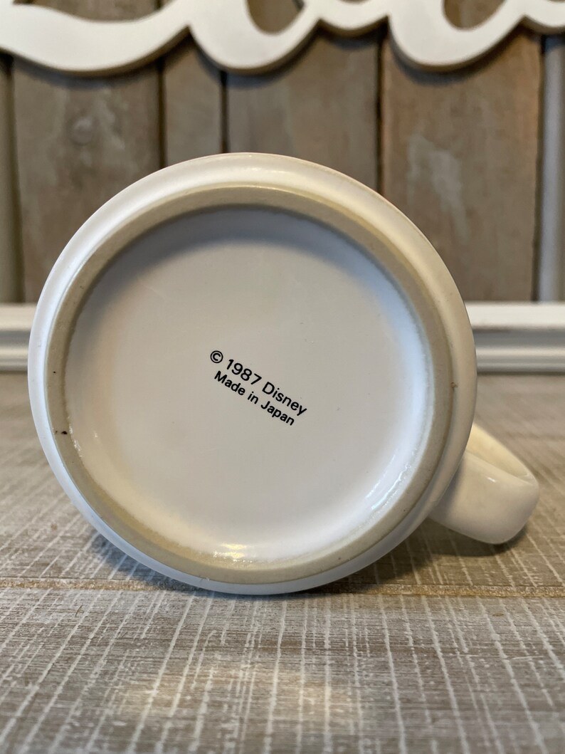 Puede incluir: Taza de cer&aacute;mica blanca con asa negra. En la parte inferior de la taza aparece el texto "&copy; 1987 Disney Hecho en Jap&oacute;n".