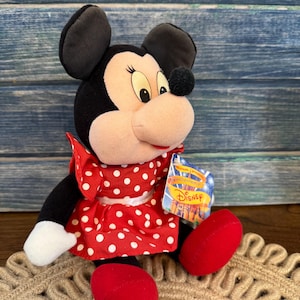 May include: A Minnie Mouse plush toy. The plush has black ears, a black head, and a light pink face. She wears a red dress with white polka dots and red shoes. A Disney tag is attached.