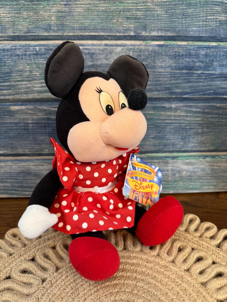 May include: A Minnie Mouse soft toy. The toy has black ears, a black head, and a light pink face. She wears a red dress with white polka dots and red shoes. A Disney tag is attached.