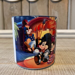 Puede incluir: Una taza de cer&aacute;mica blanca con una ilustraci&oacute;n colorida de Mickey Mouse, Minnie Mouse y Goofy frente a un edificio con el texto "Club Daisy" en el lateral.
