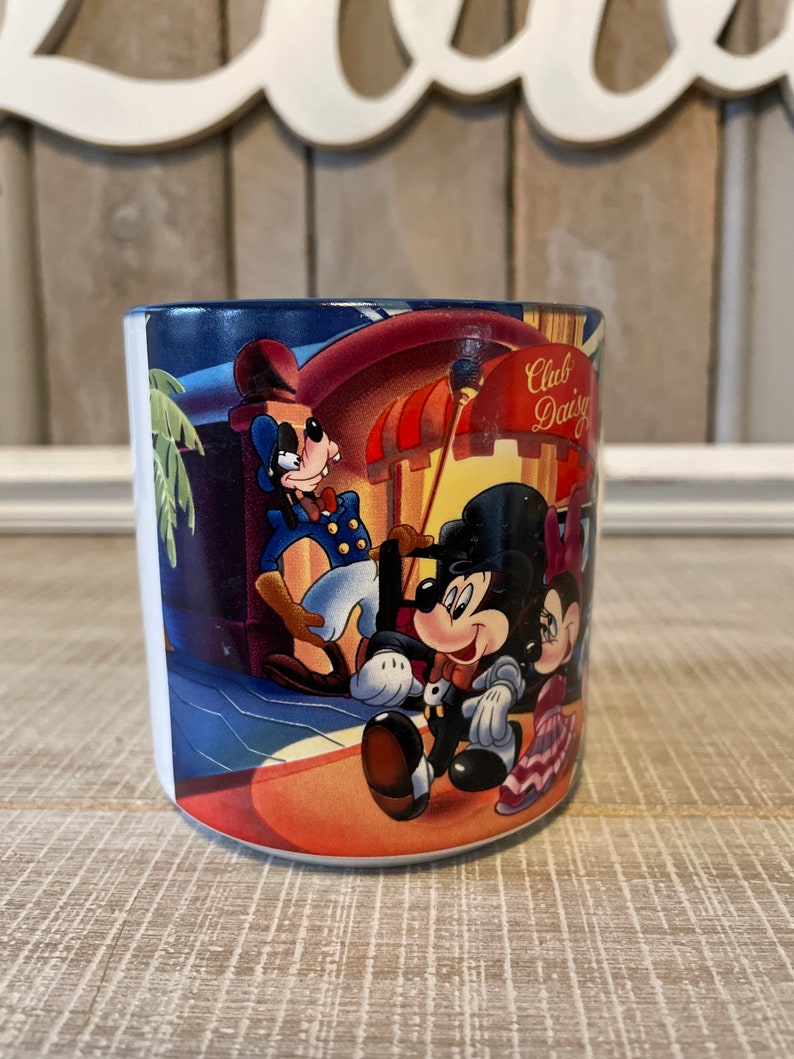 Puede incluir: Una taza de cer&aacute;mica blanca con una ilustraci&oacute;n colorida de Mickey Mouse, Minnie Mouse y Goofy frente a un edificio con el texto "Club Daisy" en el lateral.