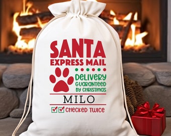 Może przedstawiać: Biały płócienny worek na prezenty od Świętego Mikołaja z czerwonym i zielonym tekstem: "Santa Express Mail Delivery Guaranteed by Christmas Milo Checked Twice". W środku worka znajduje się czerwona odbitka łapy.