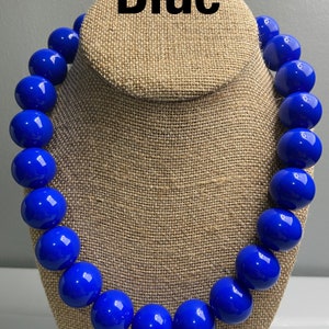 May include: A statement necklace featuring a single strand of large, round, glossy royal blue beads. The beads are uniform in size and colour, creating a bold, continuous circular design. The word Blue is printed in black text above the necklace.