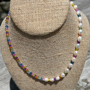 May include: A colorful beaded necklace with white pearls. The necklace is made with small, square-shaped beads in a variety of colors, including red, orange, yellow, green, blue, purple, and pink.