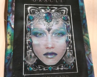 Könnte beinhalten: Eine Schachtel für ein Precious Gems Oracle Deck mit einer Frau mit einer silbernen Krone und blauen und grünen Edelsteinen. Der Hintergrund ist dunkelblau mit einem wirbelnden Muster. Der Text auf der Schachtel lautet "Precious Gems Oracle" und "Maxine Gadd."