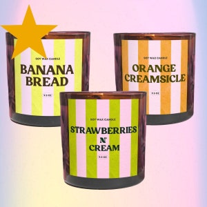 May include: Three soy wax candles with striped labels. The candles are in brown glass jars with black lids. The labels are yellow, orange, and pink with black text. The candles are labeled "Banana Bread", "Orange Creamsicle", and "Strawberries N Cream".