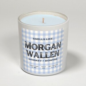 May include: A cylindrical candle in a white container with a light blue wax. The label features a blue and white gingham pattern and the text "Smells Like... MORGAN WALLEN WHISKEY + BONFIRE". The candle weighs 9.5 oz and has a burn time of 40 hours.