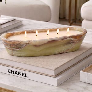 May include: An oval-shaped candle with five lit wicks, set in a green and brown stone container. The candle sits atop a stack of books, with the word "CHANEL" visible on one. The overall aesthetic is luxurious and decorative.