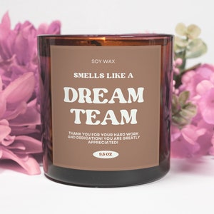 May include: A brown glass candle with a white label that reads "Soy Wax Smells Like A Dream Team Thank You For Your Hard Work And Dedication! You Are Greatly Appreciated! 9.5 OZ". The candle is surrounded by pink flowers.