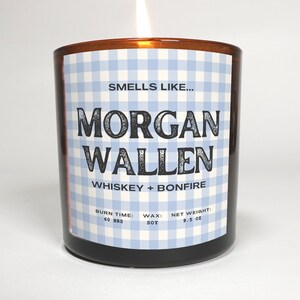 May include: A lit candle in a brown glass jar. The label features a blue and white gingham pattern and text that reads "Smells Like... Morgan Wallen Whiskey + Bonfire." The candle weighs 9.5 oz and has a burn time of 60 hours.