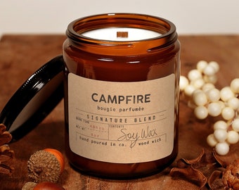 May include: A brown glass candle jar with a black lid. The candle is labeled "Campfire" and "Signature Blend" with a wood wick. The candle is surrounded by pine cones, acorns, and white berries.