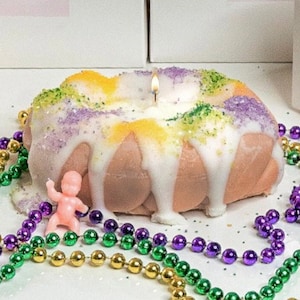 May include: A pink and white King Cake candle with purple, green, and gold sprinkles. The candle is surrounded by purple, green, and gold beads. There is a small pink baby doll on the candle.