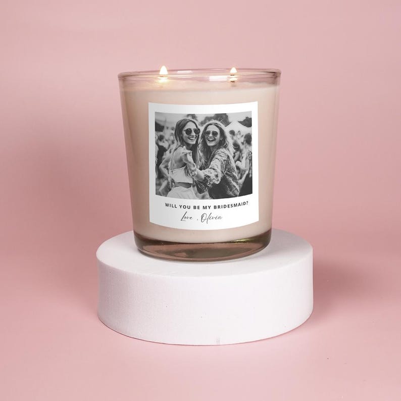 May include: A white candle with a black and white photo of two women wearing sunglasses. The text on the candle reads "Will you be my bridesmaid? Love, Olivia."