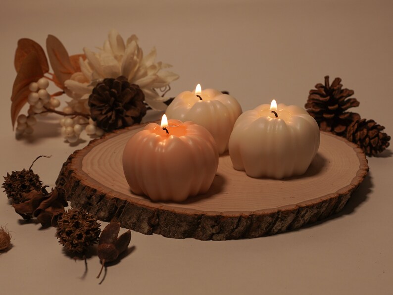 May include: Three pumpkin-shaped candles on a wooden slice. The candles are lit and are in shades of white and peach.