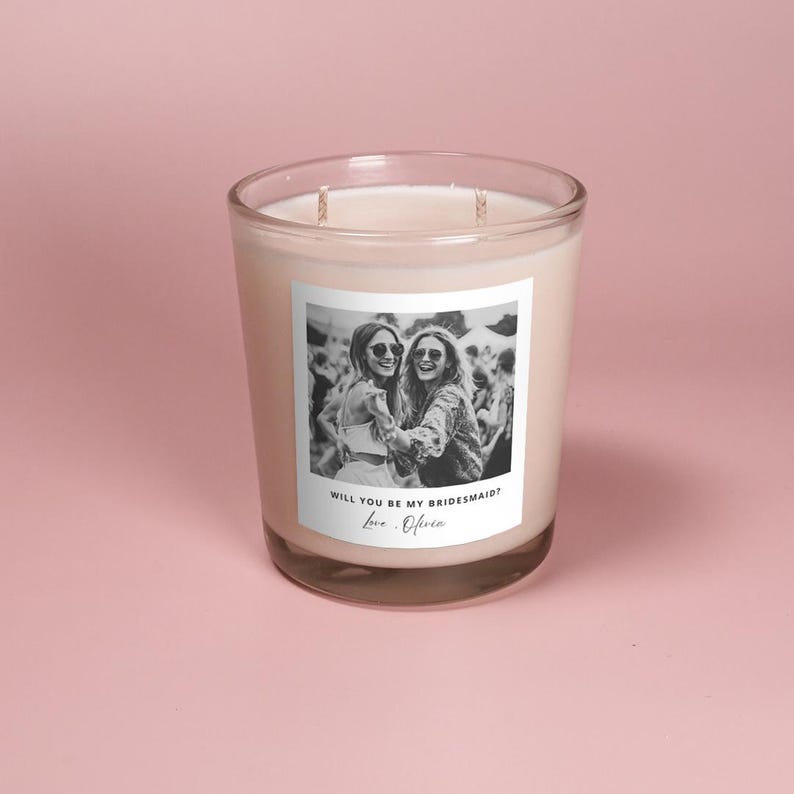 May include: A white candle with a black and white photo of two women on the label. The label reads "Will you be my bridesmaid? Love, Olivia."