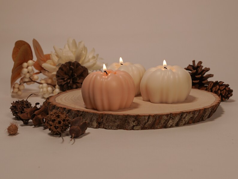 May include: Three pumpkin-shaped candles, one peach and two white, are lit and arranged on a wooden slice. The candles are surrounded by fall-themed decor, including pine cones and dried flowers.