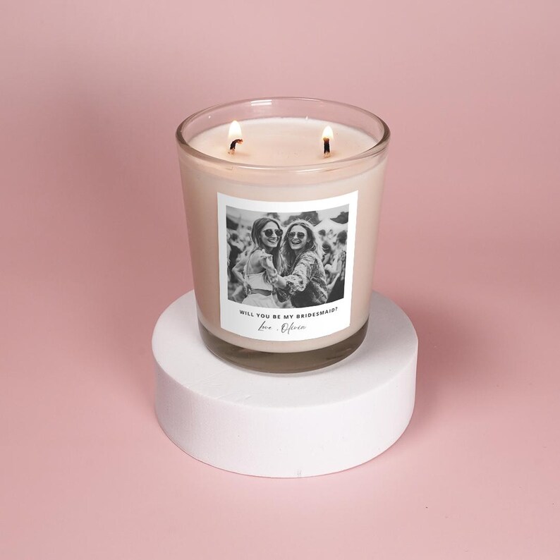 May include: A white candle with a black and white photo of two women on the front. The text on the candle reads "Will you be my bridesmaid? Love, [Name]".