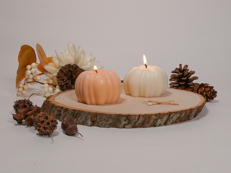 May include: Two pumpkin-shaped candles, one white and one peach, are lit and placed on a wooden slice with pine cones and fall-themed decor.
