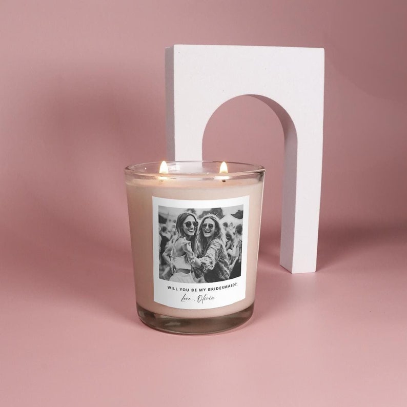 May include: A white candle with a black and white photo of two women wearing sunglasses. The text on the label reads "Will you be my bridesmaid? Love, Olivia."