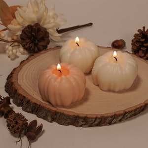 May include: Three pumpkin-shaped candles on a wooden slice. The candles are lit and are in shades of white and peach.