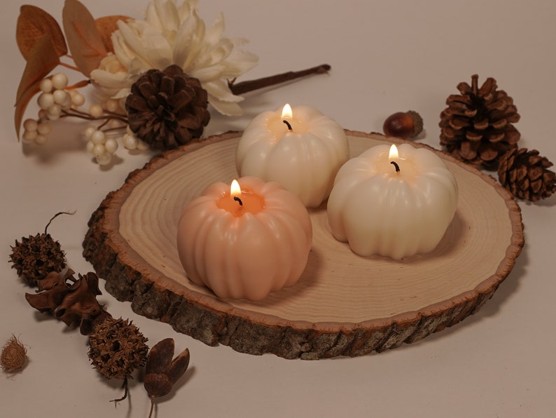 May include: Three pumpkin-shaped candles on a wooden slice. The candles are lit and are in shades of white and peach.