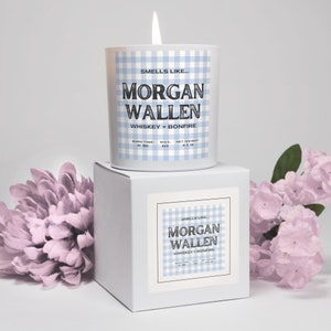 May include: A lit candle in a white glass jar with a blue gingham pattern. The label reads "Smells Like... MORGAN WALLEN WHISKEY + BONFIRE." The candle sits on a white box with a matching label. The candle weighs 9.5 oz.