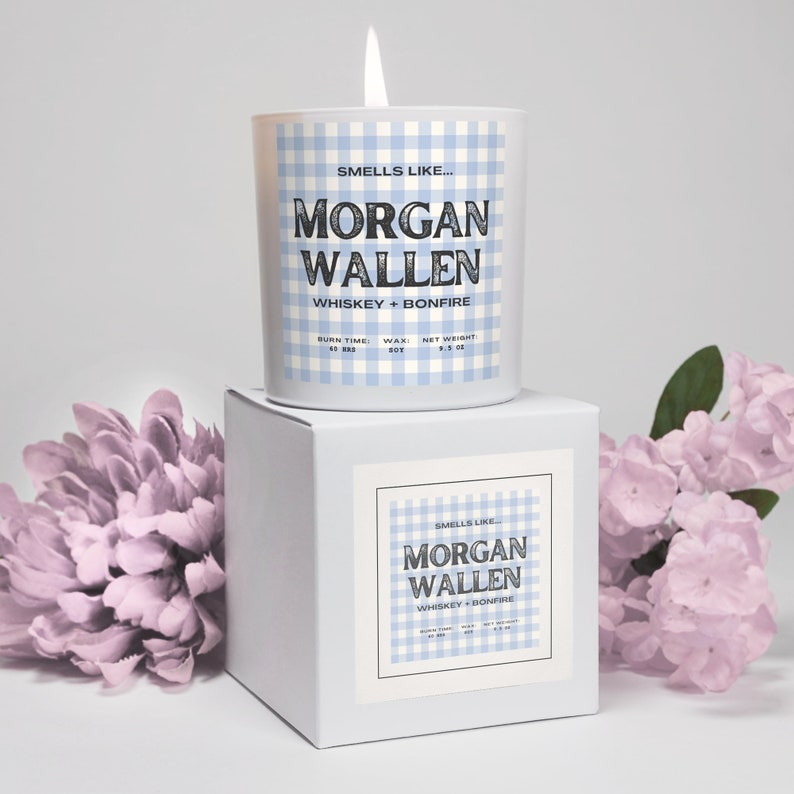 May include: A lit candle in a white glass jar with a blue gingham pattern. The label reads "Smells Like... MORGAN WALLEN WHISKEY + BONFIRE." The candle sits on a white box with a matching label. The candle weighs 9.5 oz.